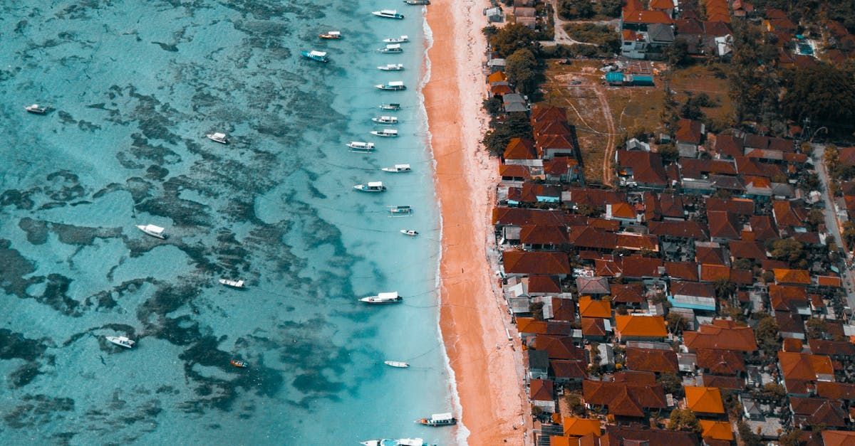 Aerial view of a beach with boats in clear blue water, sandy shore, and buildings with red roofs in Europe.