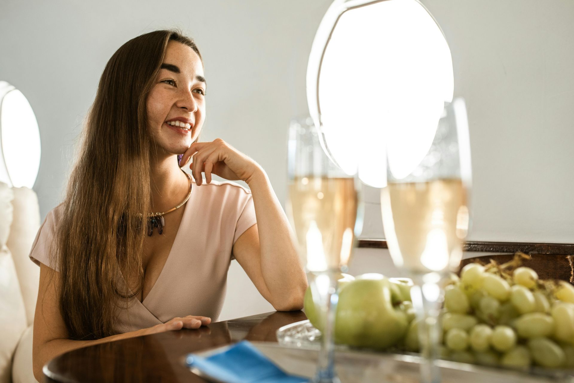 Woman smiling, hand on chin, at a table with champagne glasses and fruit on a private plane.