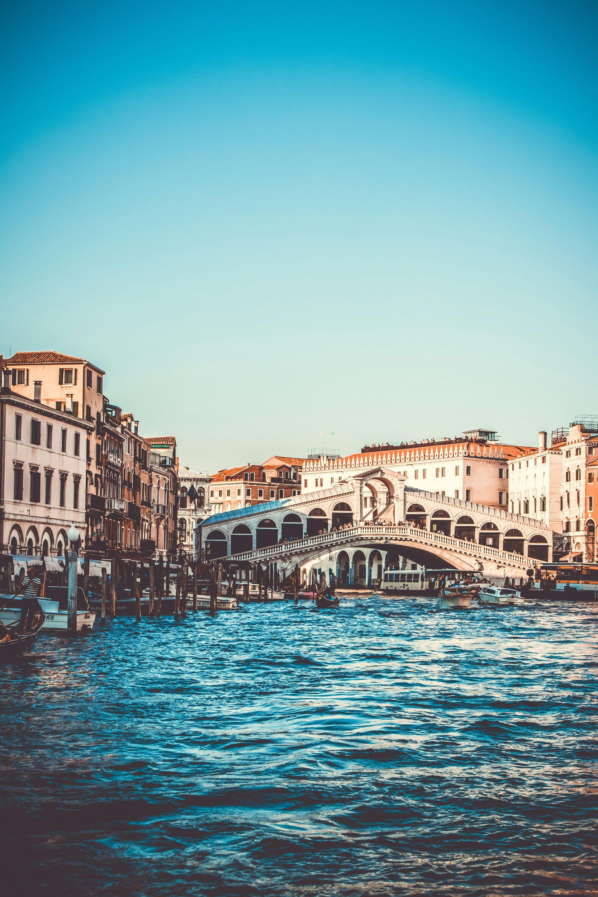 Venice cityscape with buildings lining a waterway, the Rialto Bridge in the background, blue water and sky.