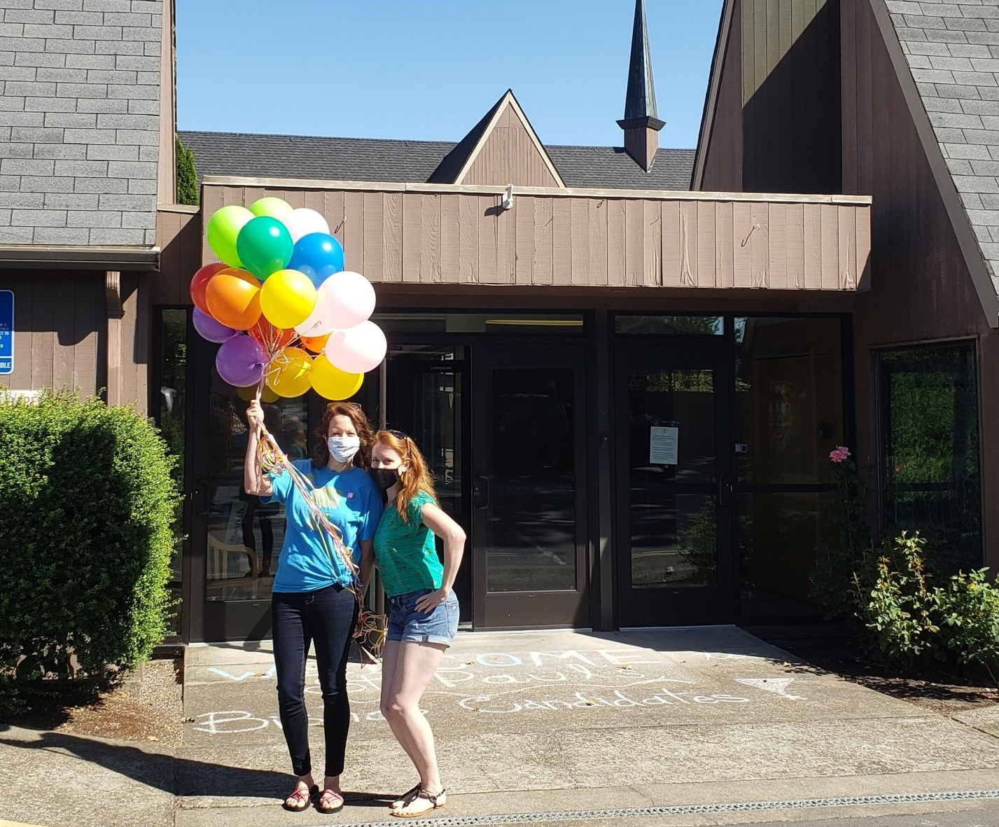 Two staff members outside of the chapel entrance at St. Paul's church. One is holding a bunch of colorful balloons, and both are wearing COVID masks.