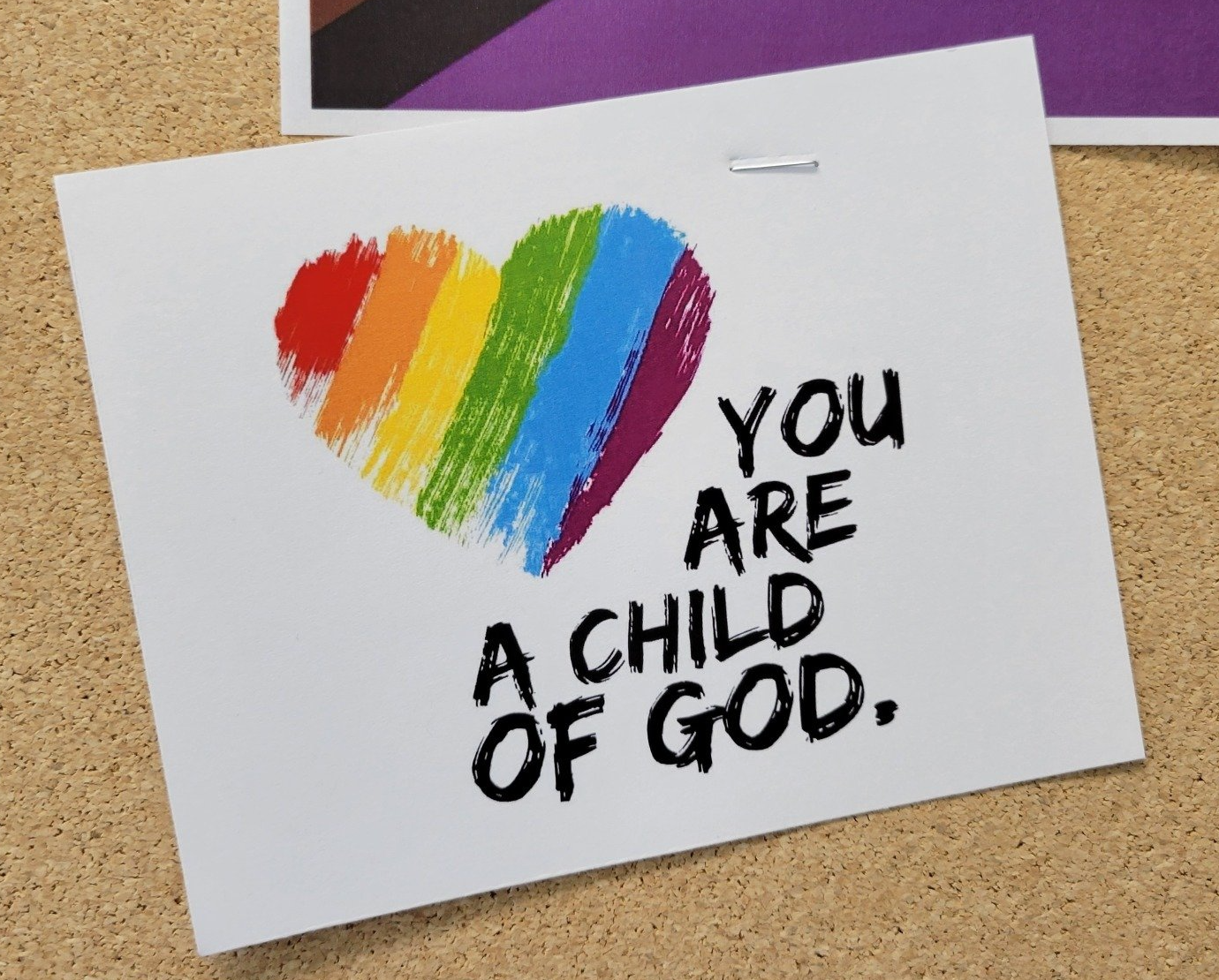 A card stapled to a cork board. The card is white with a rainbow heart and text reading 