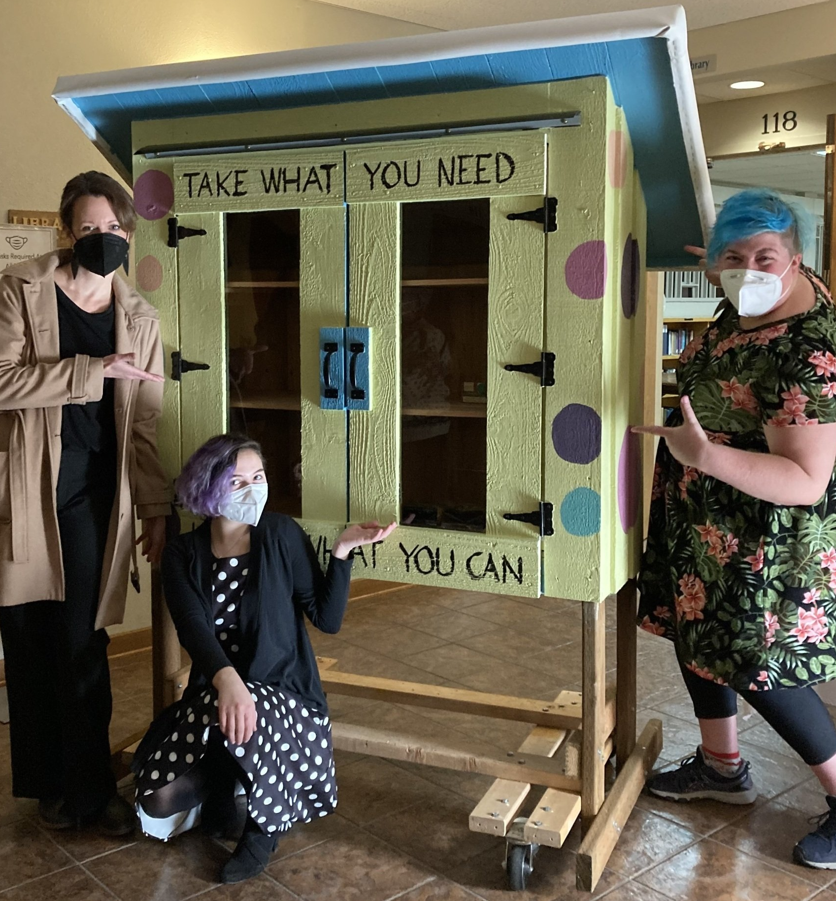 Three happy humans pointing at the newly completed Dot Pantry as it rests in the lobby, waiting to be blessed. The pantry is painted yellow with different colored polkadots and text reading 