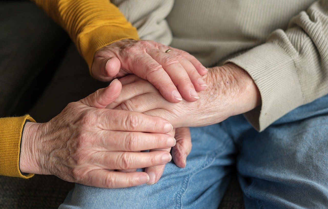 An elderly couple is holding hands while sitting on a couch.