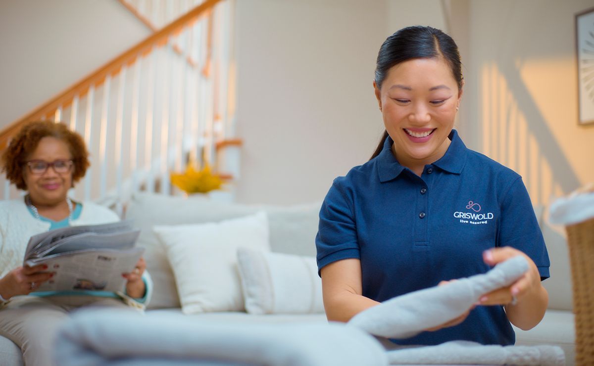 A woman is folding a blanket in a living room while another woman sits on a couch reading a newspaper.