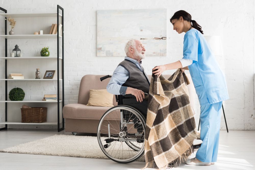 Caregiver assisting a person in a wheelchair with a blanket in a living room.