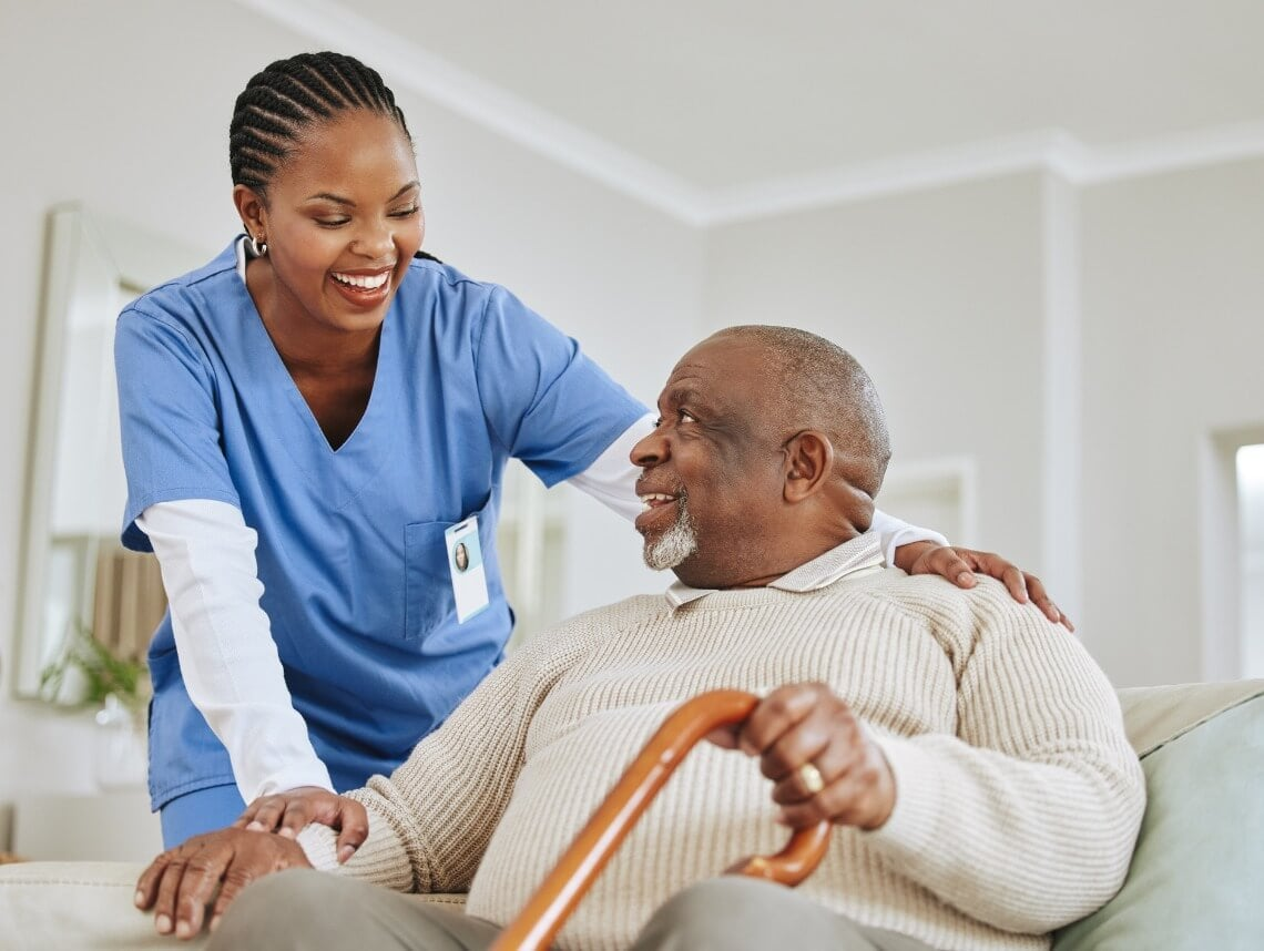 Caregiver assisting a person seated on a couch; both smiling, inside a home.