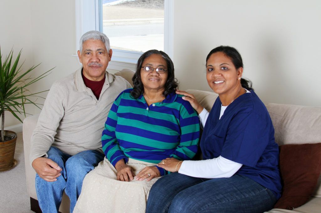 An older couple sits on a couch with a caregiver; they smile indoors.