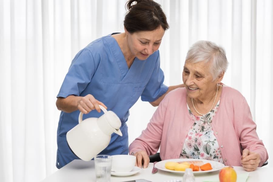 Caregiver pouring liquid for a senior woman at a dining table.