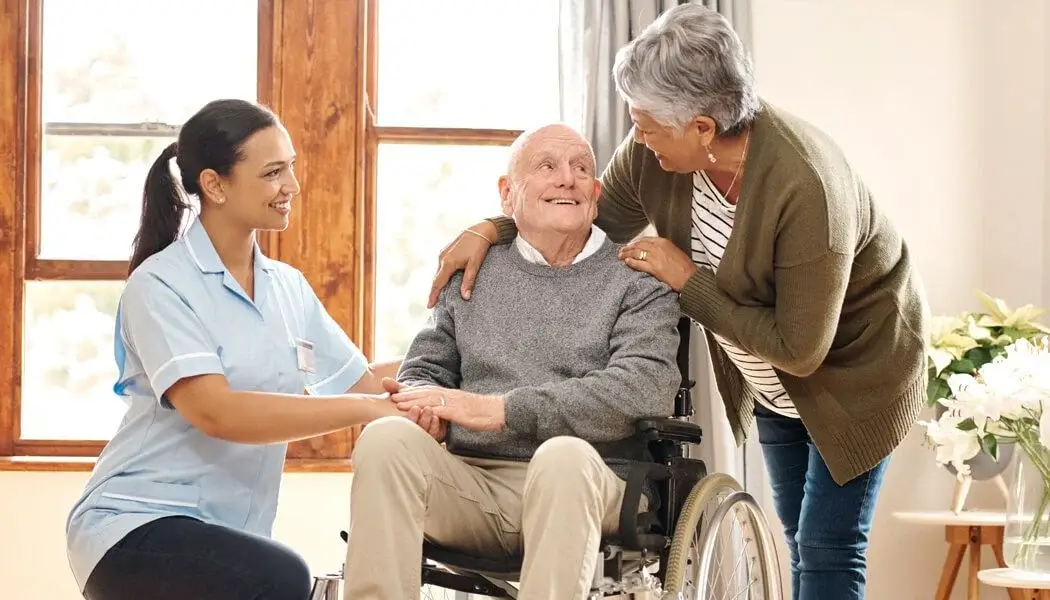 Woman in scrubs with an elderly man in a wheelchair and another woman, all smiling in a home setting.