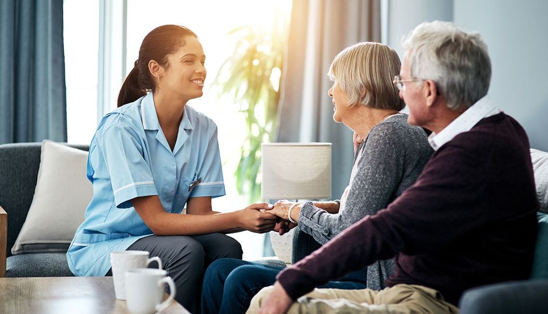 A nurse is holding the hands of an elderly couple while they sit on a couch.