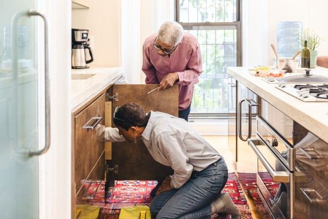 Two people unpacking boxes in a bright kitchen, one kneeling under the sink and one standing by the counter.