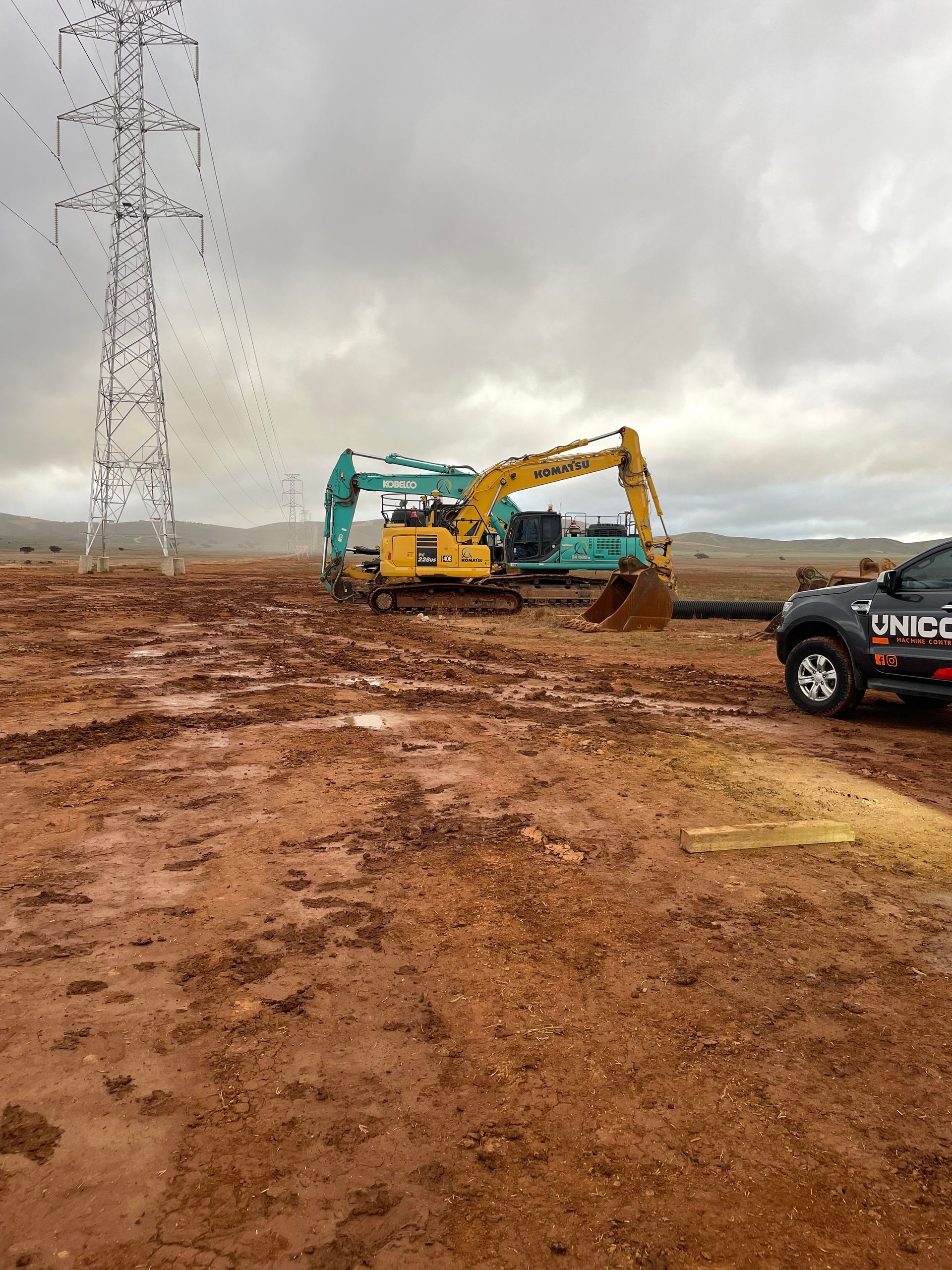 Two excavators on muddy ground near power lines on an overcast day. A vehicle is on the right.
