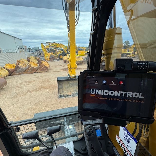 View from inside an excavator cab, displaying a UNICONTROL screen, with other yellow construction equipment visible outside.