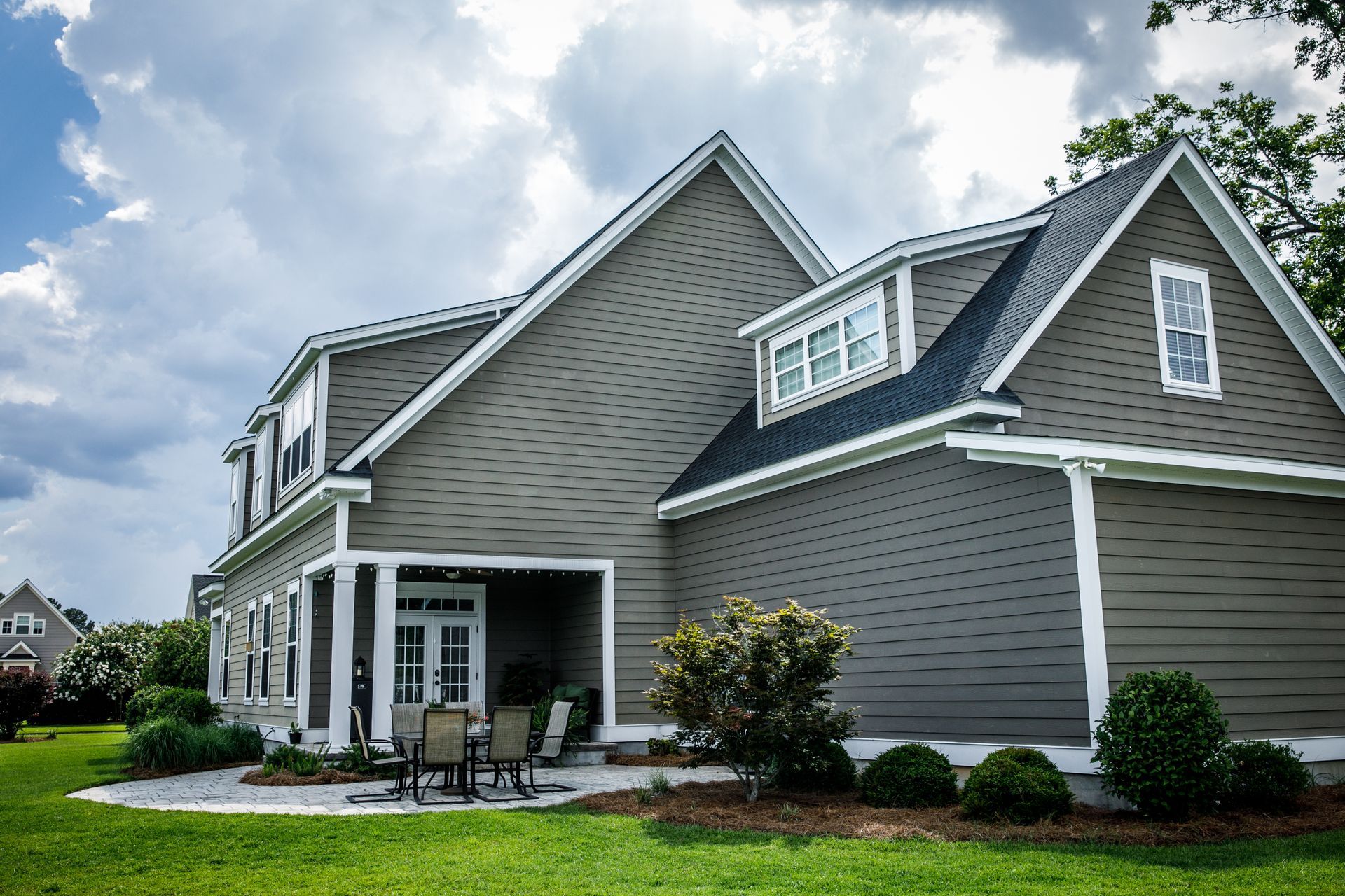 Two-story house with gray siding, dormers, and a small patio, set in a grassy yard under a cloudy sky.