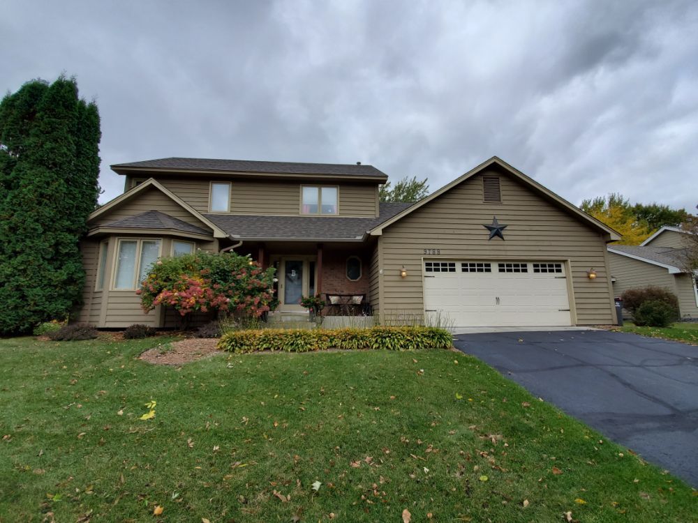 Two-story tan house with a garage and green lawn under a cloudy sky.