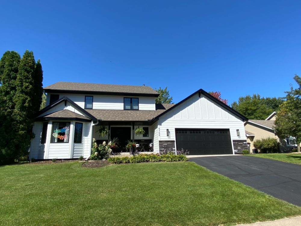 White two-story house with black trim, garage door, and a bay window, on a green lawn under a blue sky.