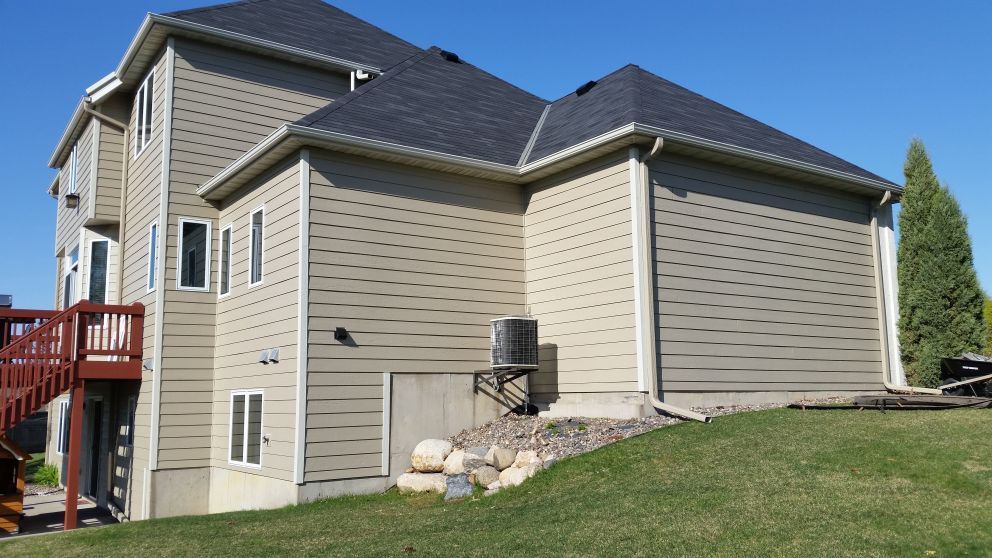 Two-story house with beige siding, dark roof, and a deck. Located on a grassy hill under a blue sky.