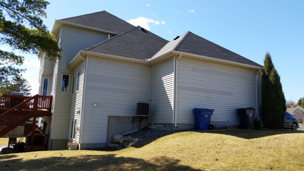 Side view of a two-story beige house with a brown deck, blue bins, and a grassy yard under a blue sky.