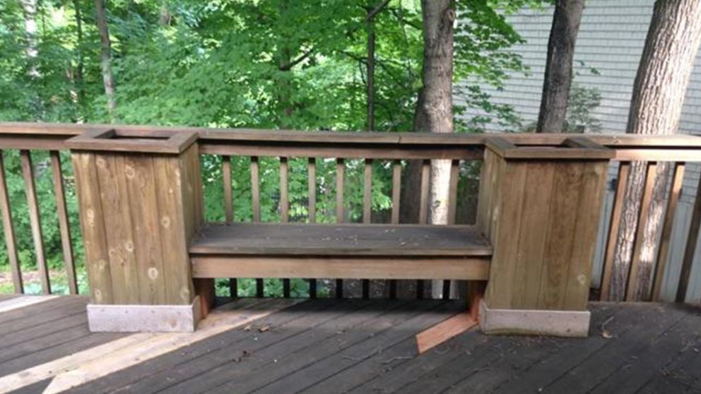 Wooden bench on a deck, flanked by planters, with a forest background.