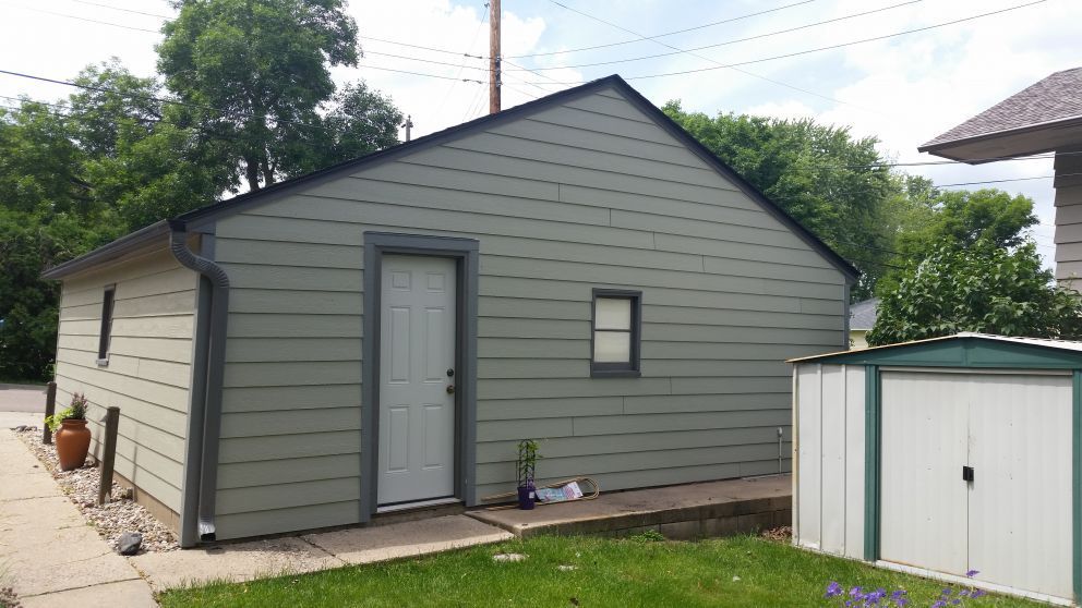 Green detached garage with a white door and small window, next to a shed.