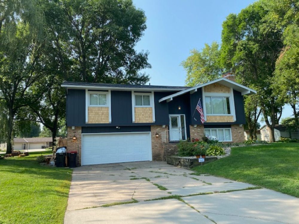 Two-story house with dark blue siding, beige trim, and a white garage door. Trees surround the home on a sunny day.