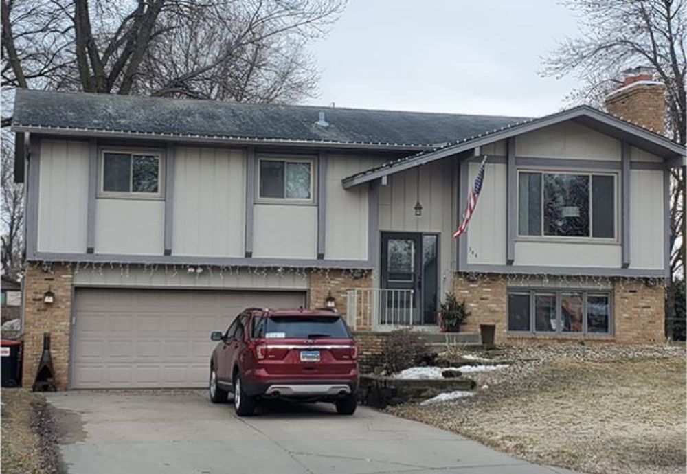 Two-story beige house with red SUV in driveway, American flag hanging, gray roof, brick accents.