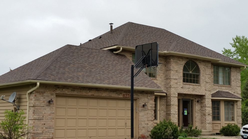 Two-story brick house with brown roof and garage, basketball hoop in front. Overcast sky.