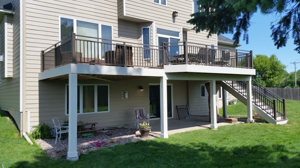 Two-story house with a deck. The deck has black railings, with outdoor furniture on both levels. Beige siding, green grass.