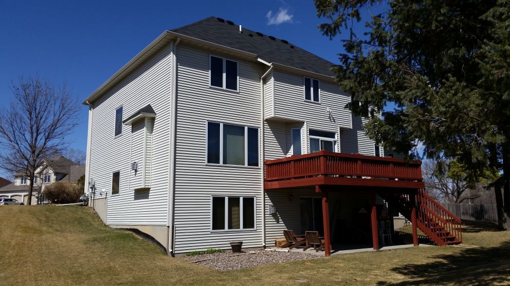 Two-story white house with a brown deck and stairs, set on a grassy hill under a blue sky.