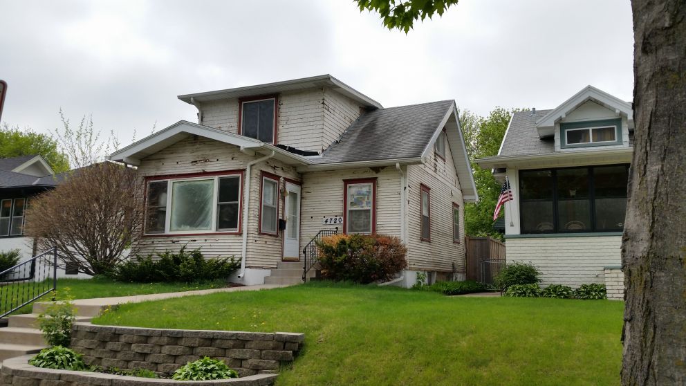 Two-story house with weathered siding and red trim on a green lawn, adjacent house on the right. Overcast sky.