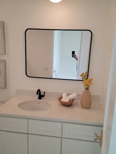 Bathroom with white cabinets, light countertop, black-framed mirror, and vase of yellow flowers.