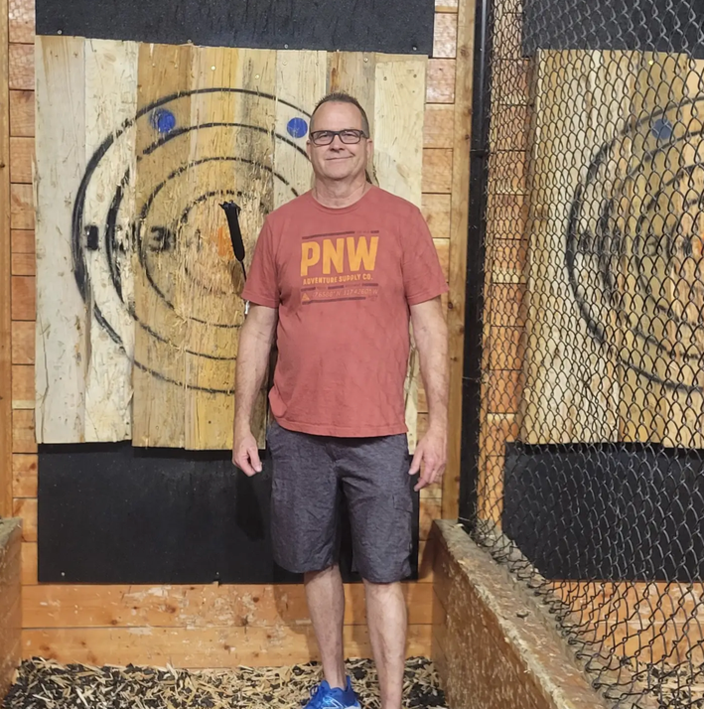 Man stands in front of axe throwing targets. He wears a red shirt, grey shorts, and blue shoes. Wooden setting.