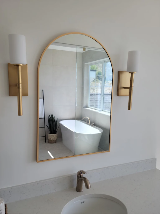 Bathroom with gold-framed arch mirror, two sconces, and a white soaking tub reflected in the mirror.
