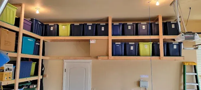 Garage shelving unit filled with storage bins, mainly blue, yellow, and green, above a white door and garage door opener.