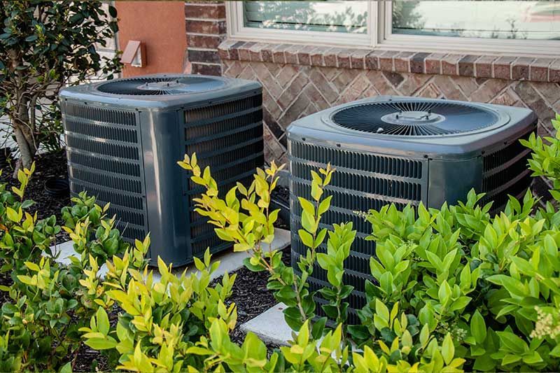 Two outdoor air conditioning units beside a house, partially surrounded by green shrubs.