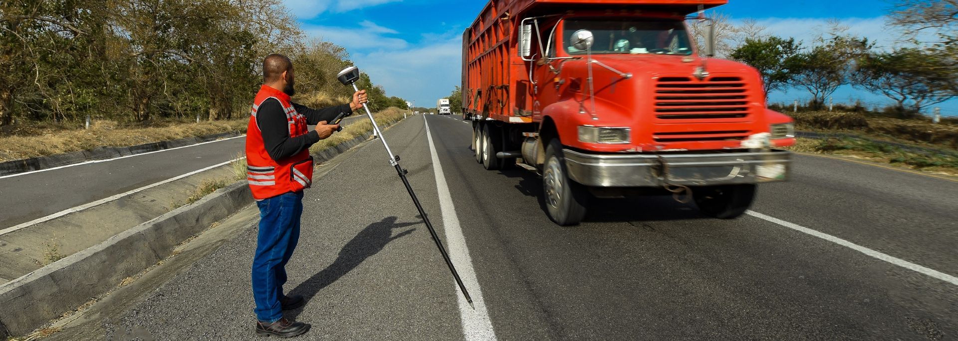 Um homem está parado na beira de uma rodovia ao lado de um caminhão vermelho.