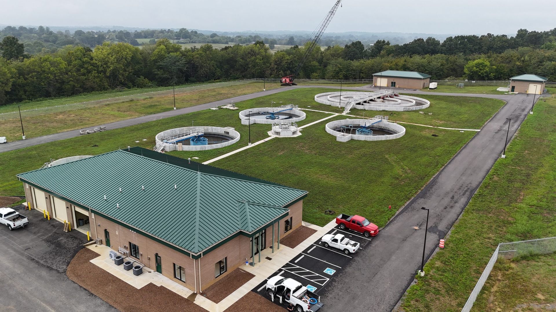 Aerial view of a wastewater treatment plant with several circular tanks and a long building; a truck is parked in front of the building.