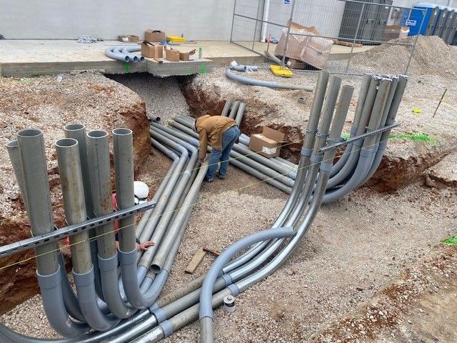 Construction worker installs gray electrical conduits in a trench. Gravel, dirt, and equipment surround him.