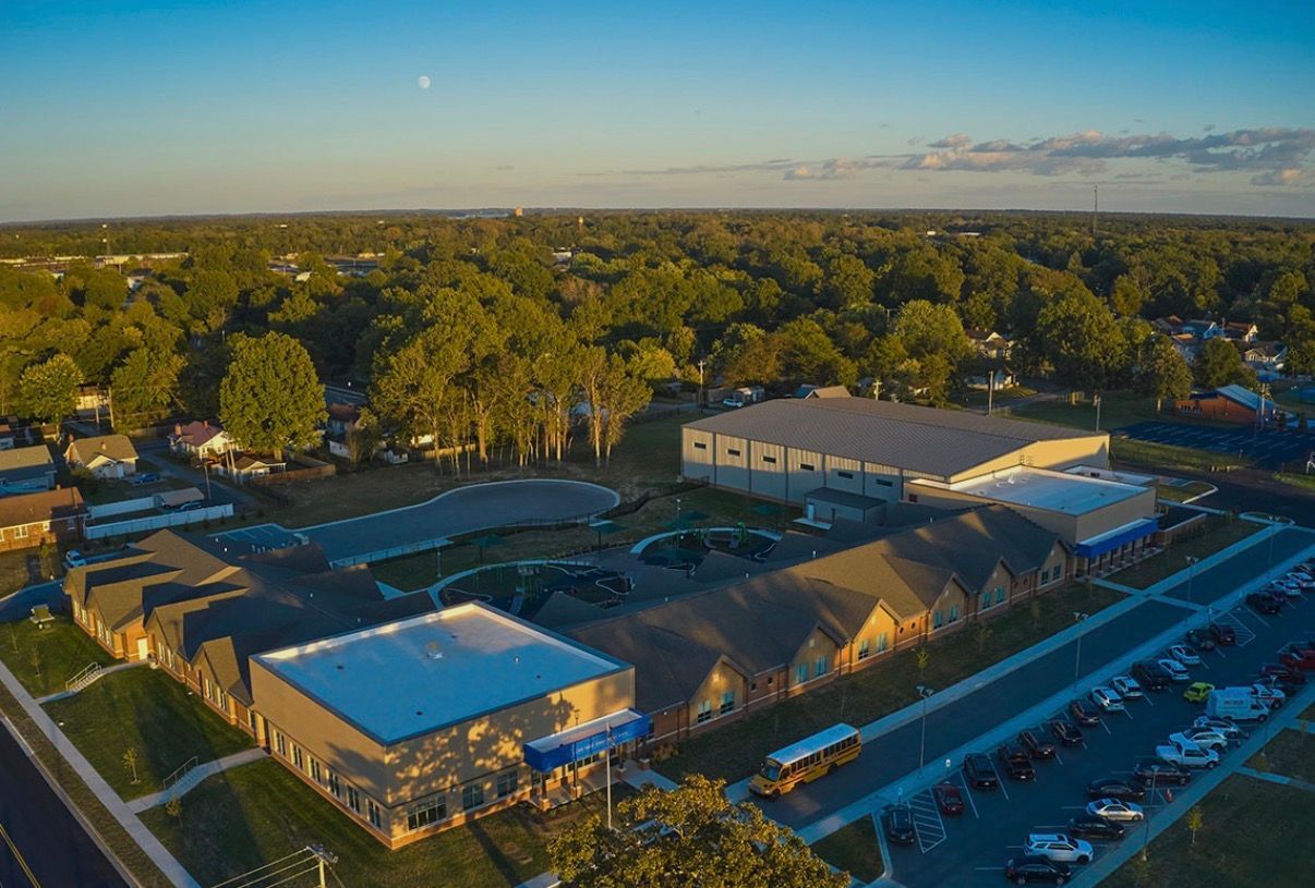 Aerial view of a school building with surrounding trees and a parking lot, under a blue sky.