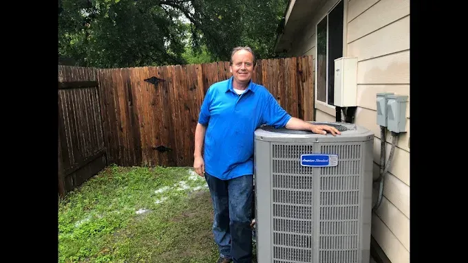 A man in a blue shirt is standing next to an air conditioner on the side of a house.