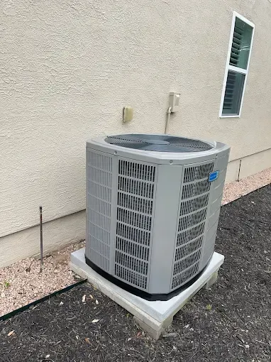 A large air conditioner is sitting outside of a house next to a window.
