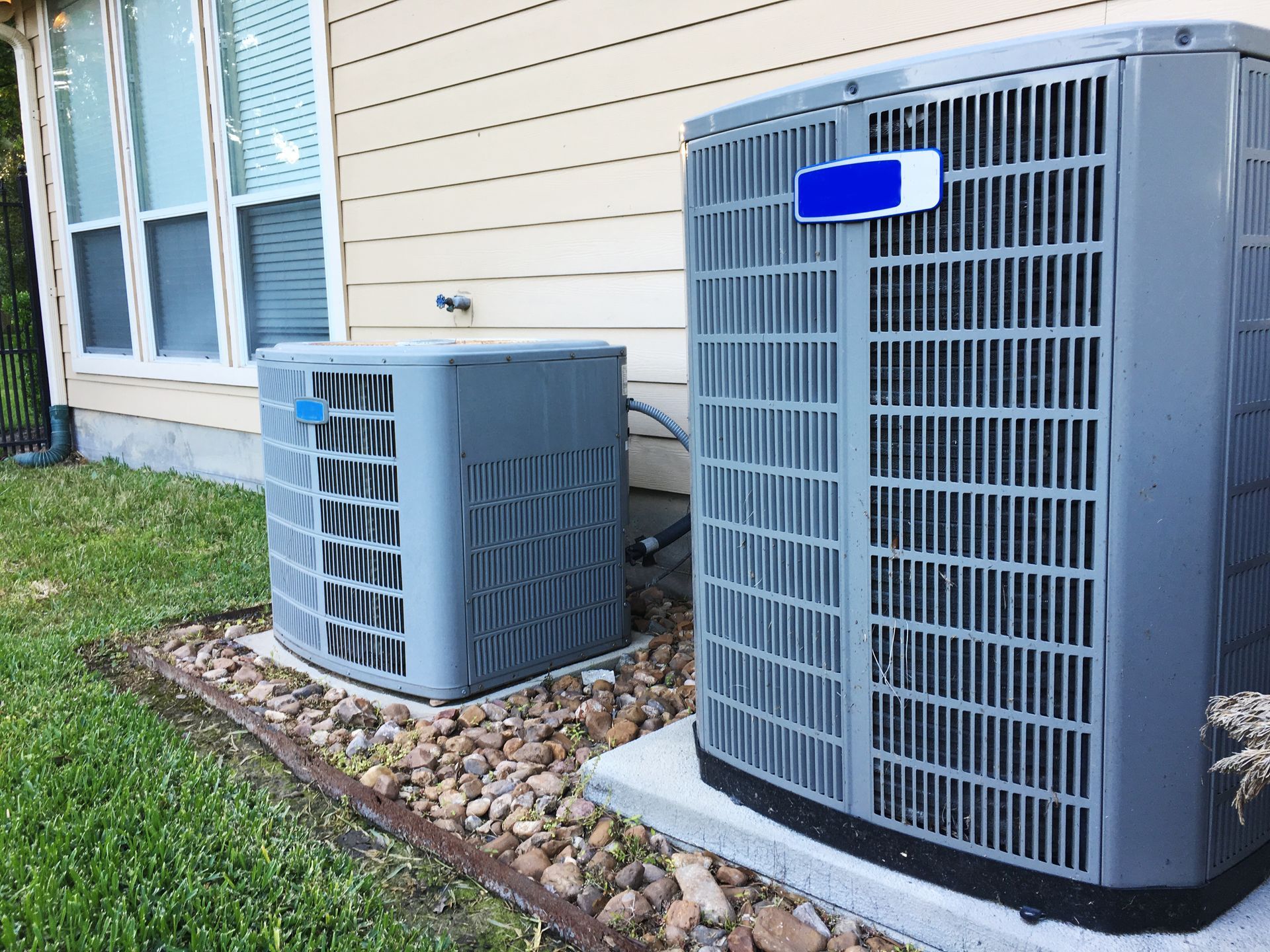 Two gray air conditioning units outside a building with a window, set on gravel.