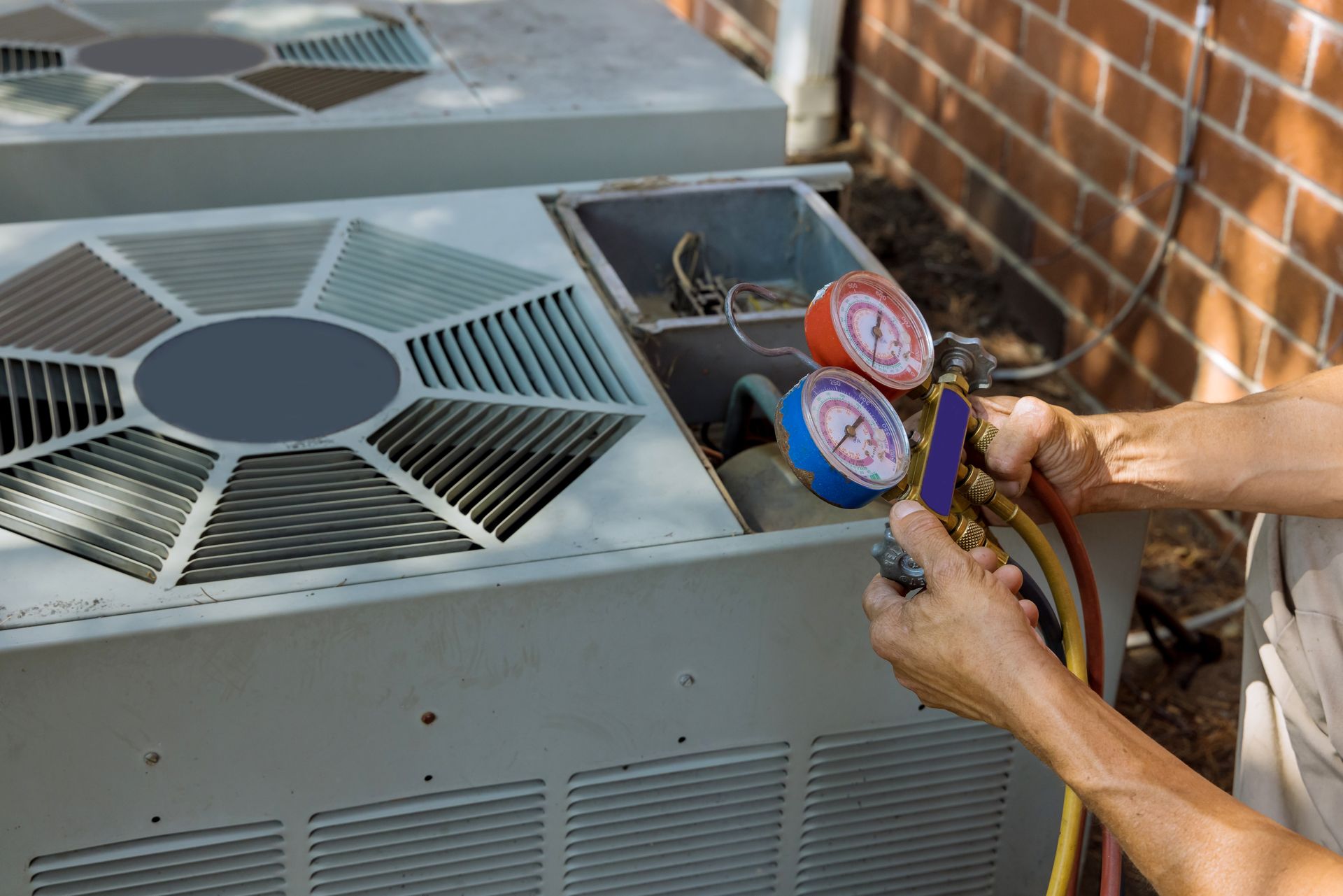 Person servicing an air conditioning unit with gauges, outdoors near a brick wall.