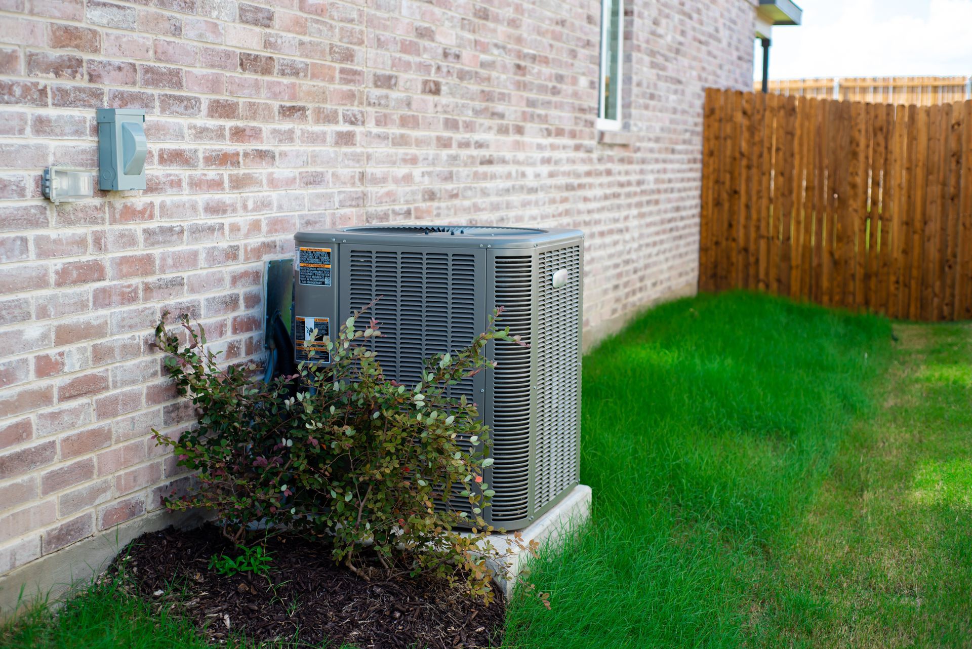 Air conditioning unit next to a brick wall and wooden fence, set in a grassy yard.