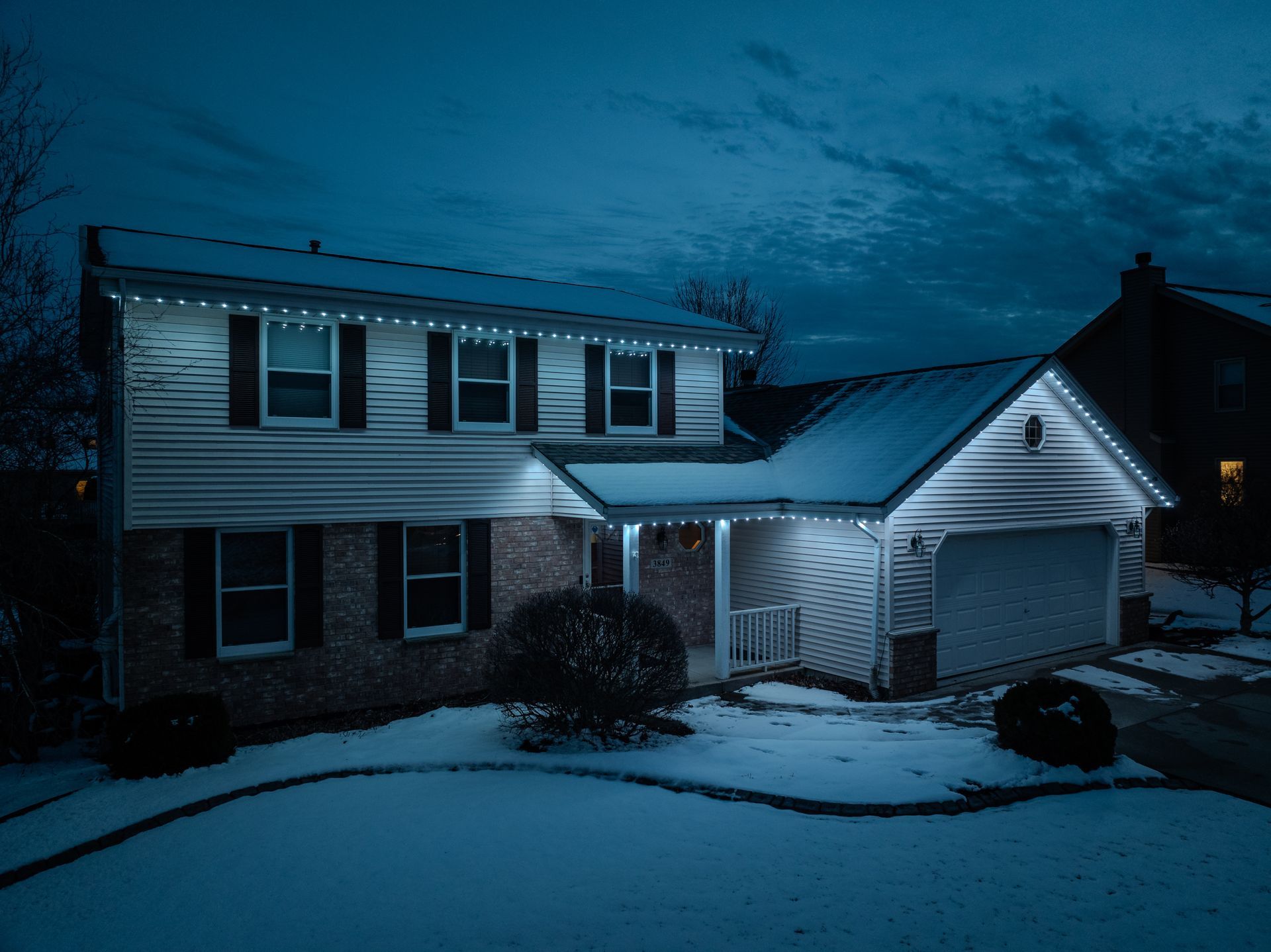 A house with pretty white lights on it is covered in snow at night.
