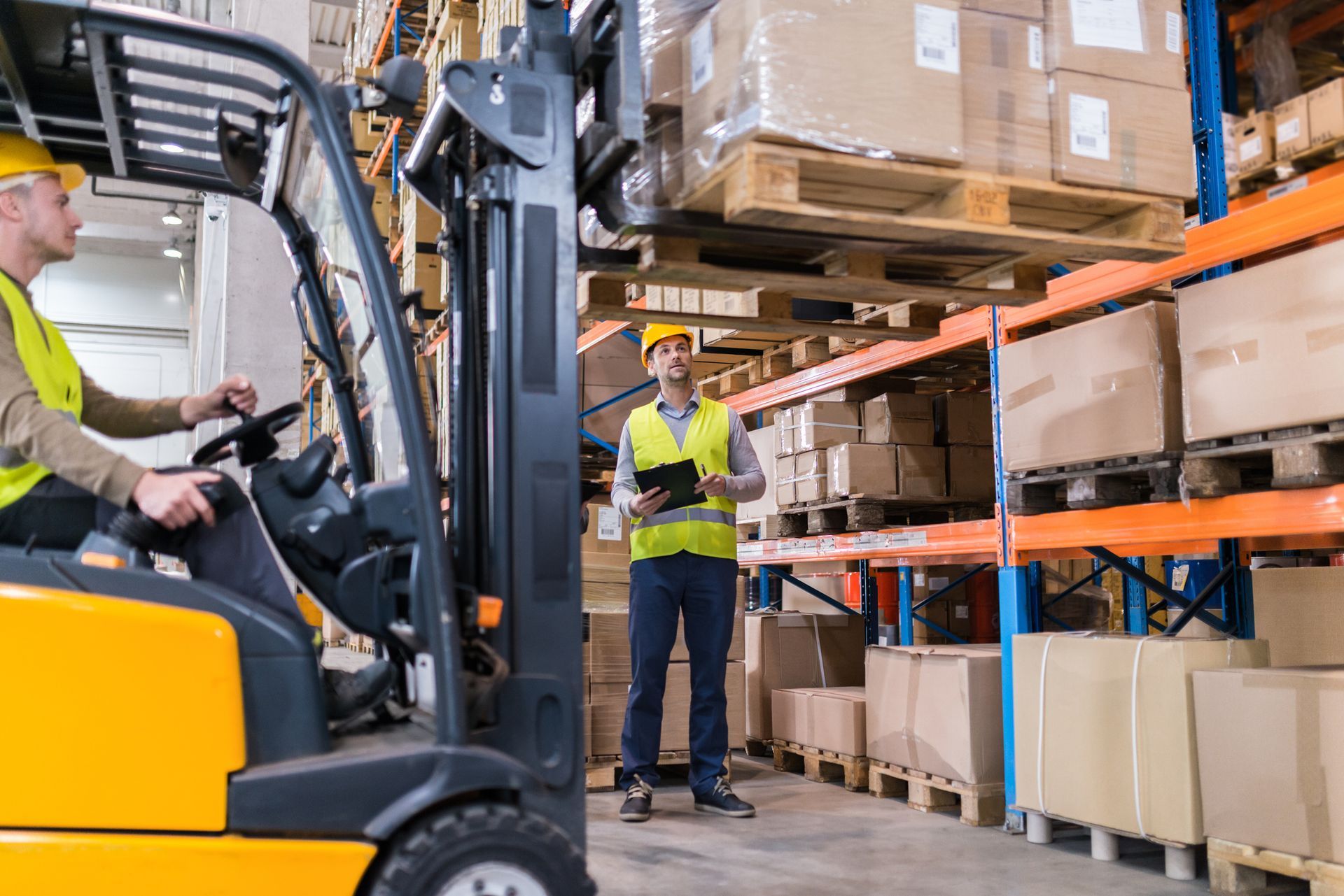 A man is driving a forklift in a warehouse while another man looks on.