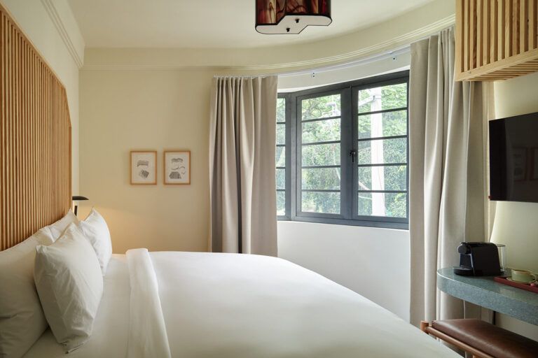 Habitación de hotel con cama, ventana, cortinas y obras de arte. Tonos beige y neutros con luz natural.