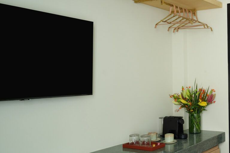Televisor de pantalla plana en una pared blanca sobre un mostrador con cafetera, flores y perchas.