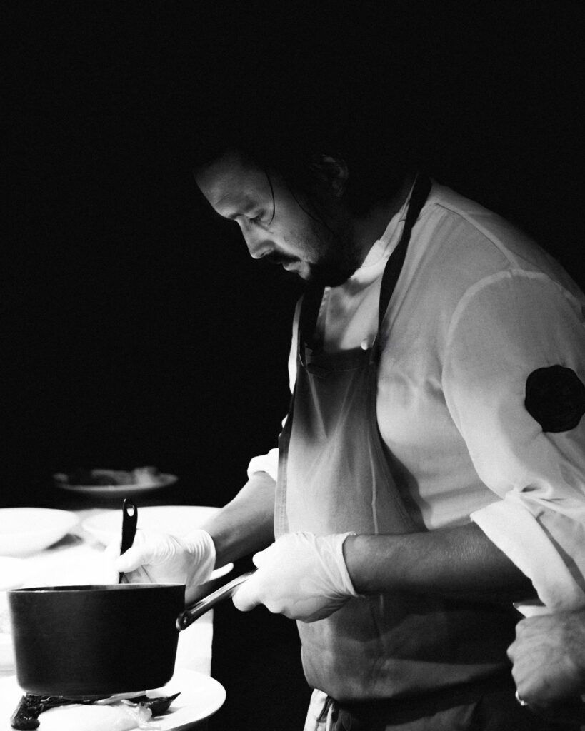 Un chef con bata blanca y delantal revuelve una olla en una cocina poco iluminada.