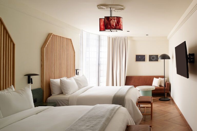 Habitación de hotel con dos camas, un sofá y un televisor montado en la pared; tonos neutros con una lámpara decorativa.